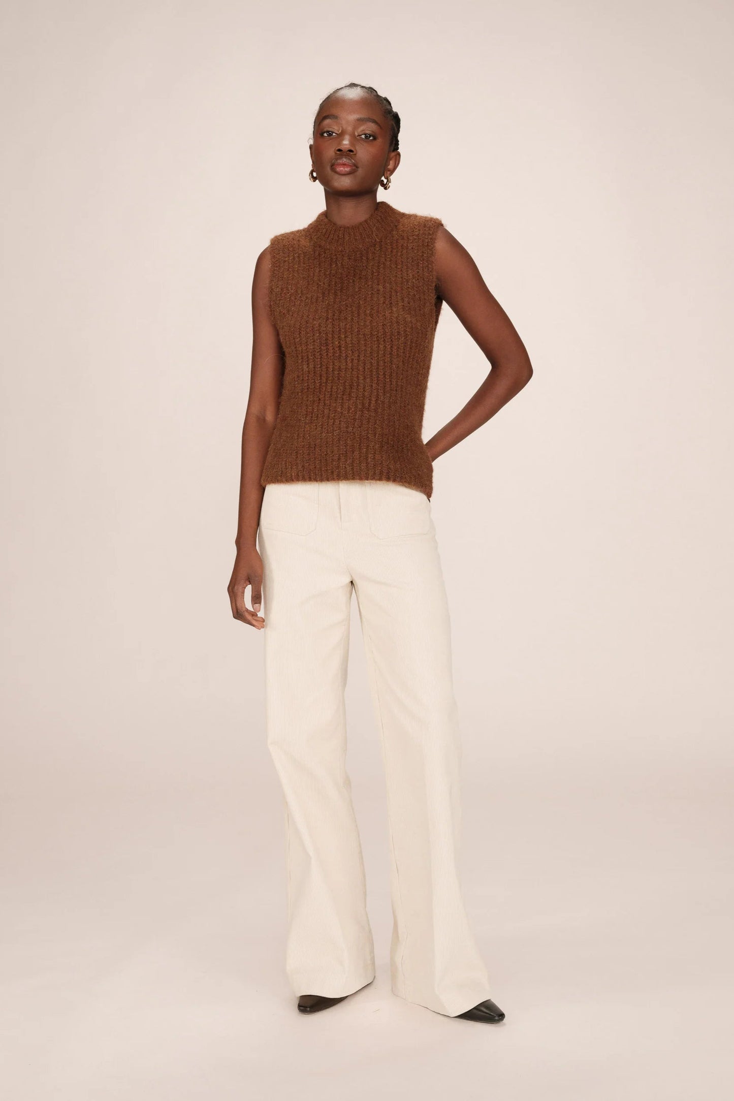 Woman wearing a brown sleeveless sweater and white pants on a beige background