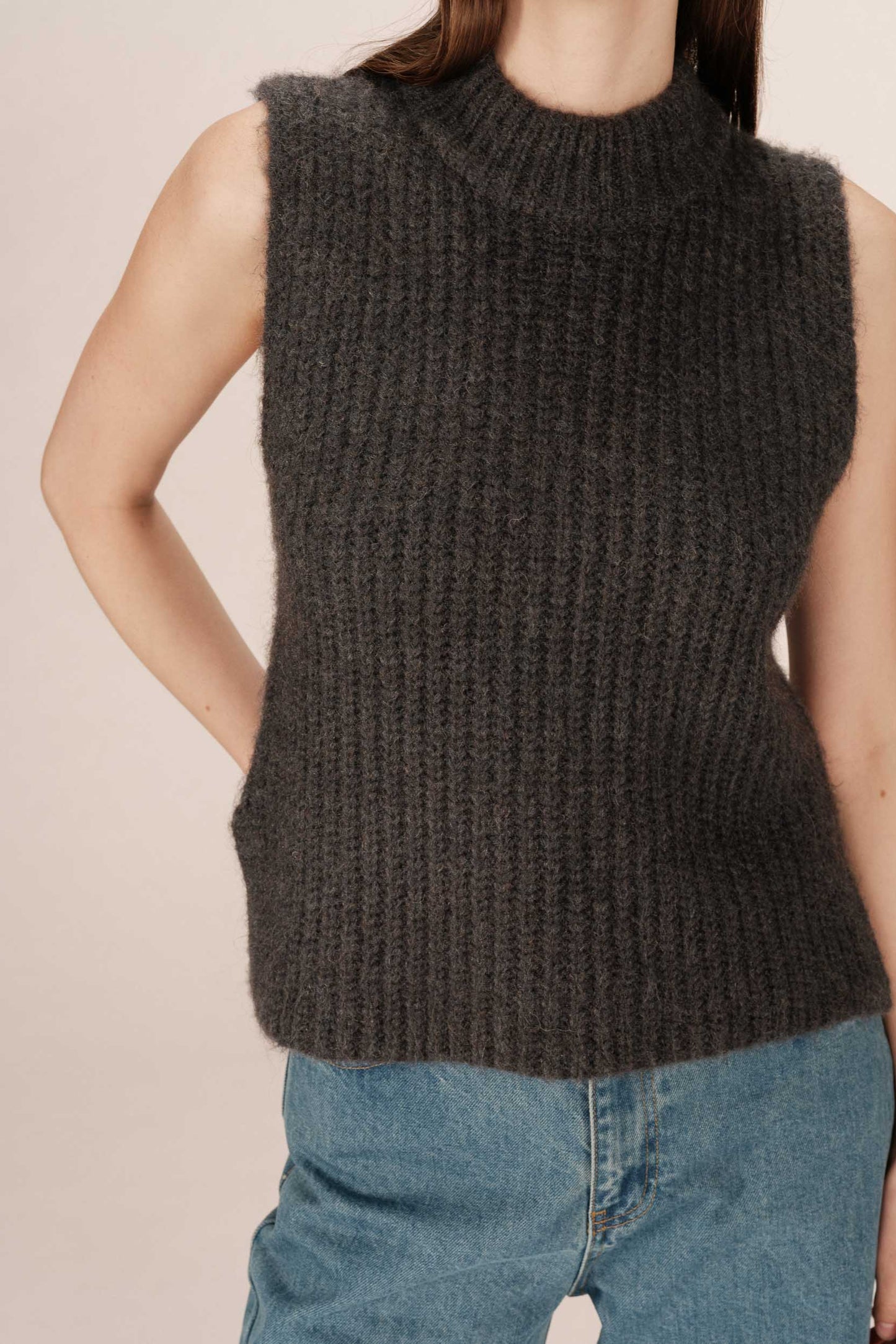 Person wearing a dark brown sleeveless knitted sweater with blue jeans on a beige background