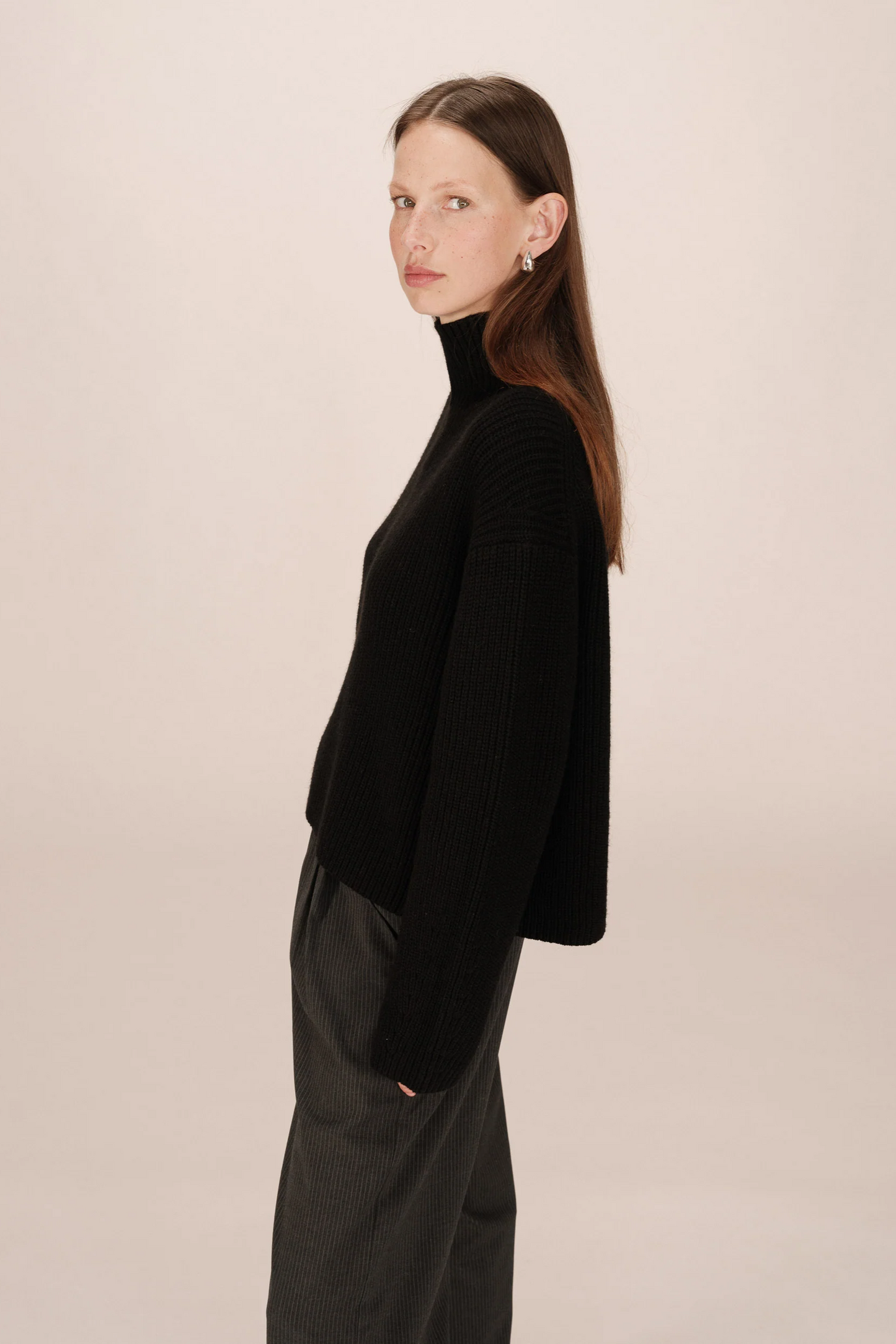 Woman wearing a black turtleneck sweater and dark pants against a light pink background
