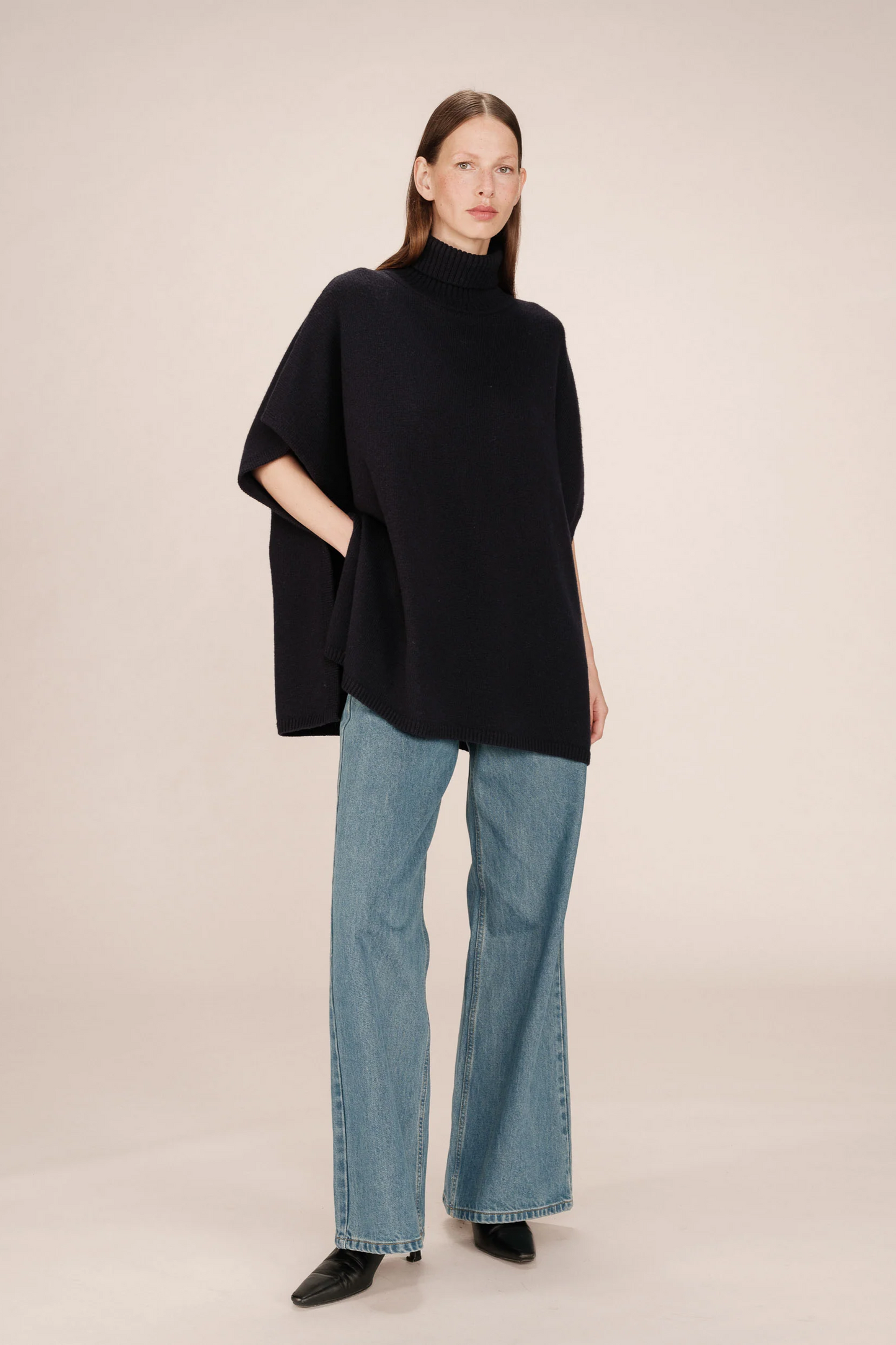 Person wearing a black poncho and blue jeans on a beige background