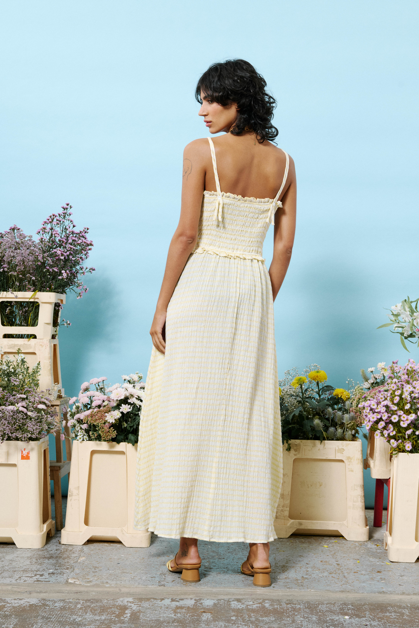 Woman in a butter yellow dress standing among potted plants with a blue sky background