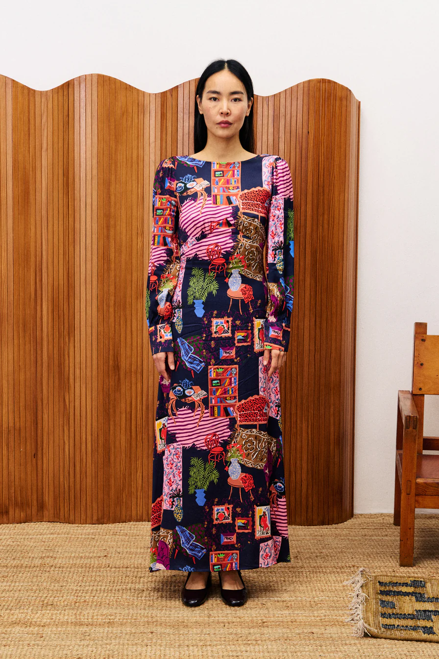 Woman wearing a long, patterned dress standing in front of a wooden screen.