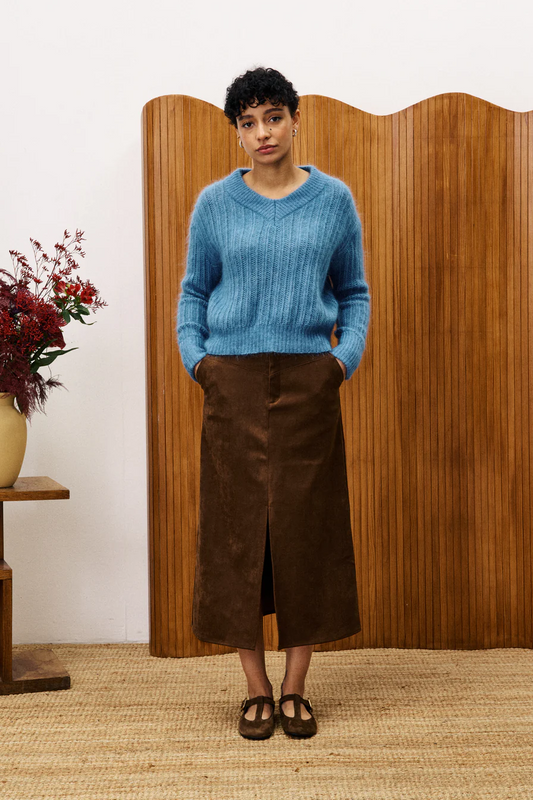 Person wearing a blue sweater and brown skirt standing in front of a wooden screen.