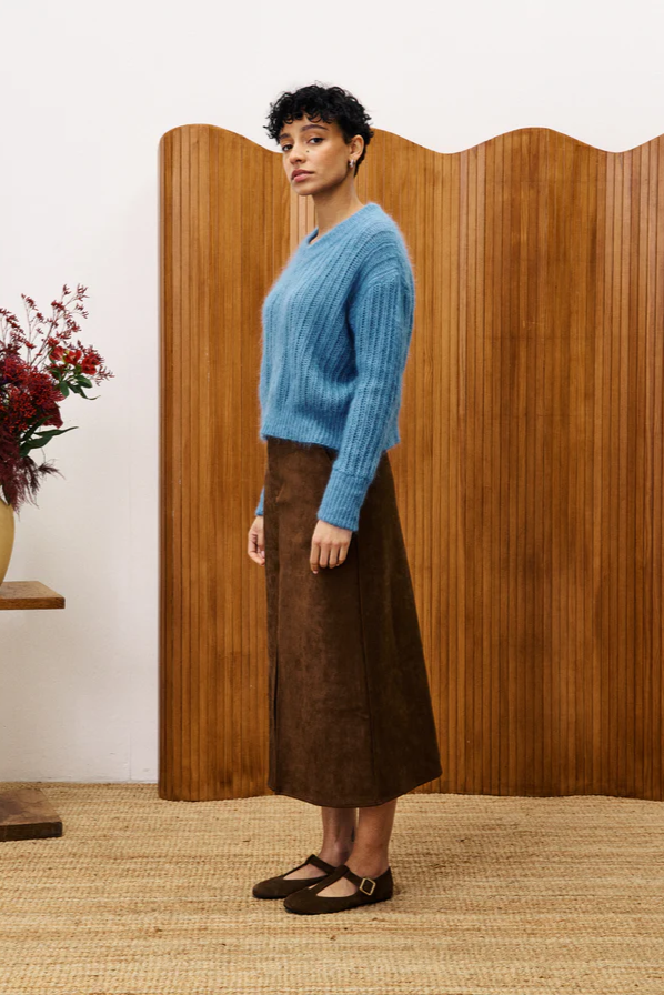 Woman wearing a blue sweater and brown skirt standing in front of a wooden screen.