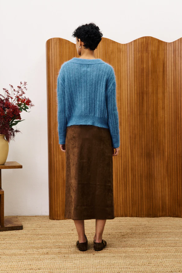 Person wearing a blue sweater and brown skirt standing in front of a wooden screen.