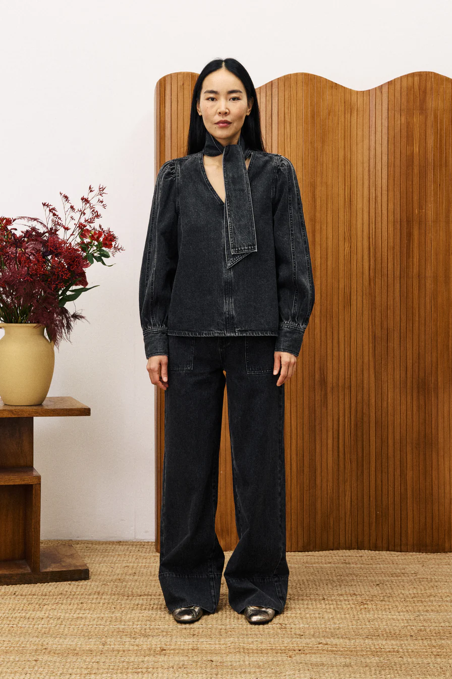 Person wearing a black denim outfit standing in front of a wooden screen with a vase of flowers on a table.