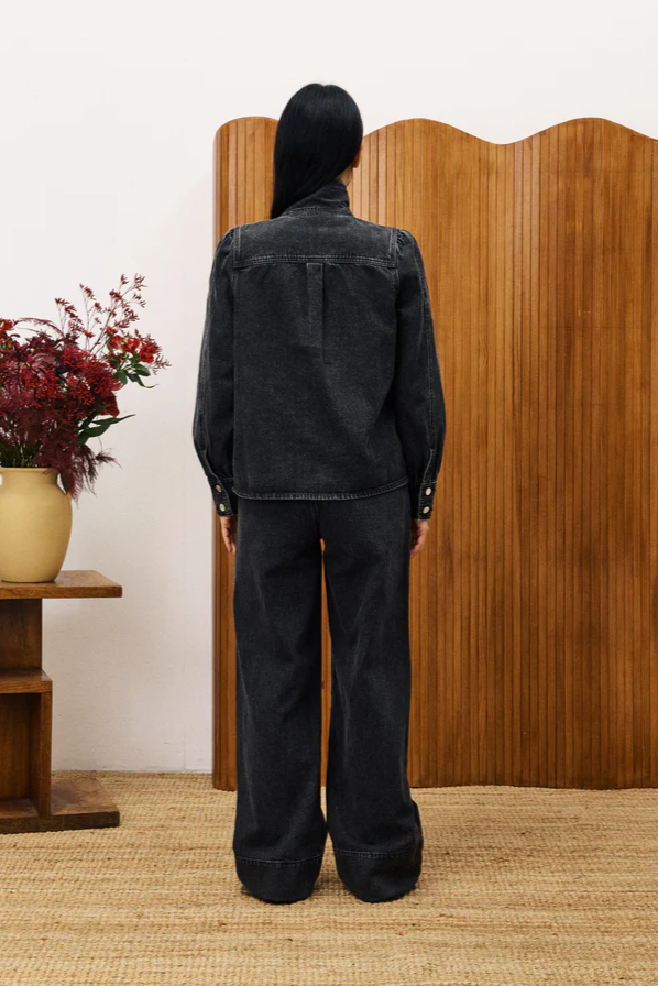 Person wearing a black denim jacket and pants standing in front of a wooden screen with a vase of flowers on a table.