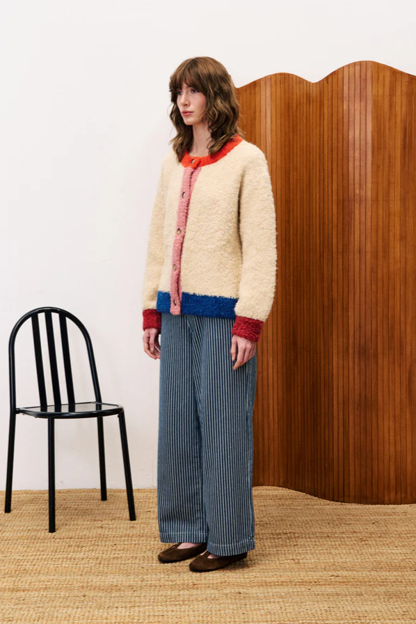 Woman wearing a cream cardigan with red trim and blue pants standing against a white wall with a wooden panel.