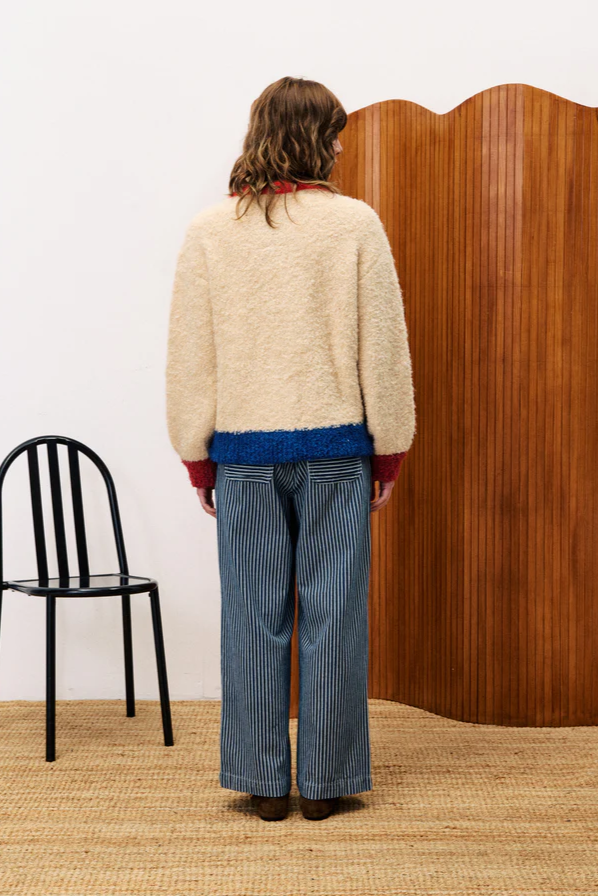 Person wearing a cream sweater and blue pants standing in front of a wooden screen with a black chair on a beige carpet.