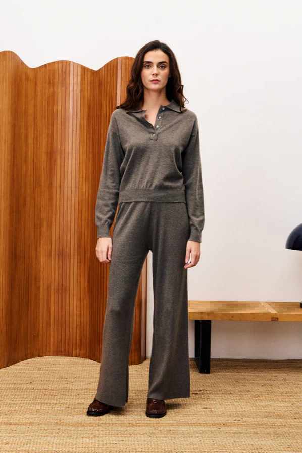 Woman wearing a gray outfit standing in a room with wooden paneling and a lamp.