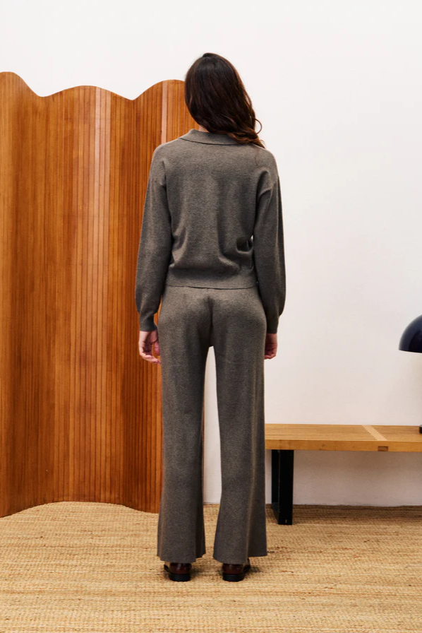 Person wearing a gray suit standing in a room with wooden paneling and a lamp.