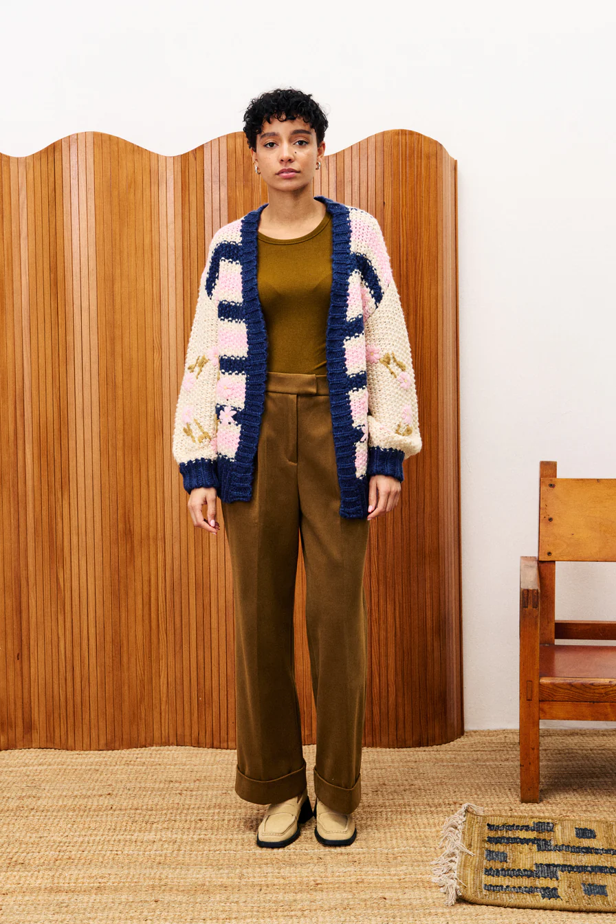 Person wearing a colorful knitted cardigan, brown top, and pants standing in front of a wooden screen.