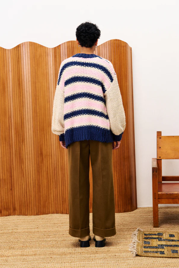 Person wearing a colorful striped sweater and brown pants standing in a room with wooden furniture.