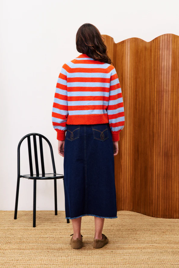 Person wearing a red and white striped sweater and blue skirt standing in a room with a black chair and wooden screen.