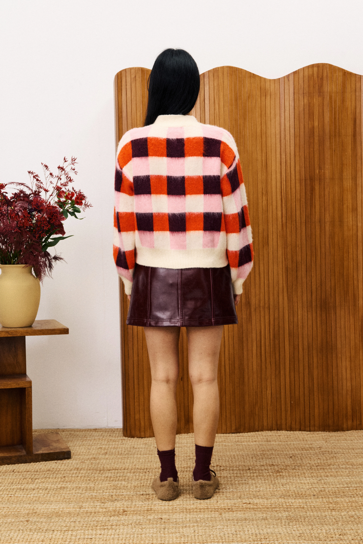 Person wearing a checkered sweater and leather skirt standing in front of a wooden screen with a vase of flowers on a table.