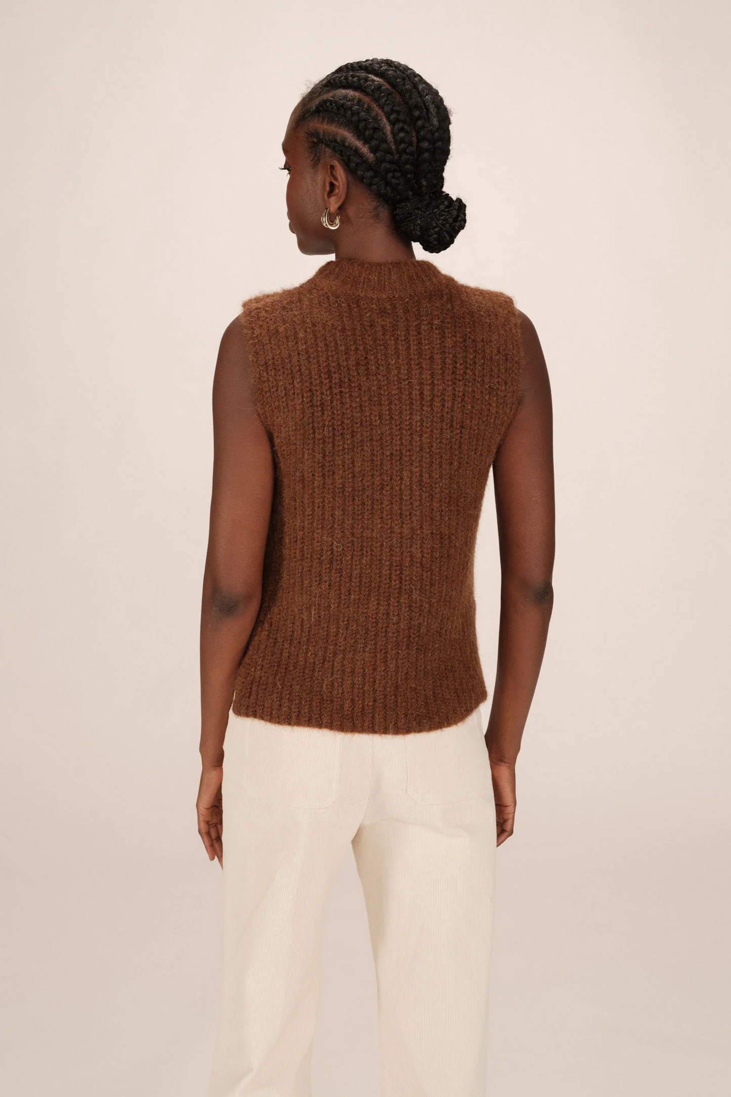 Person wearing a brown knitted vest and white pants on a beige background