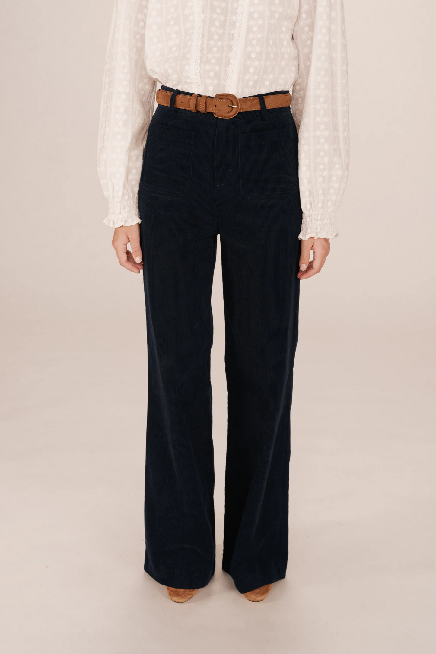 Person wearing a white blouse and dark blue velvet pants with a brown belt on a beige background