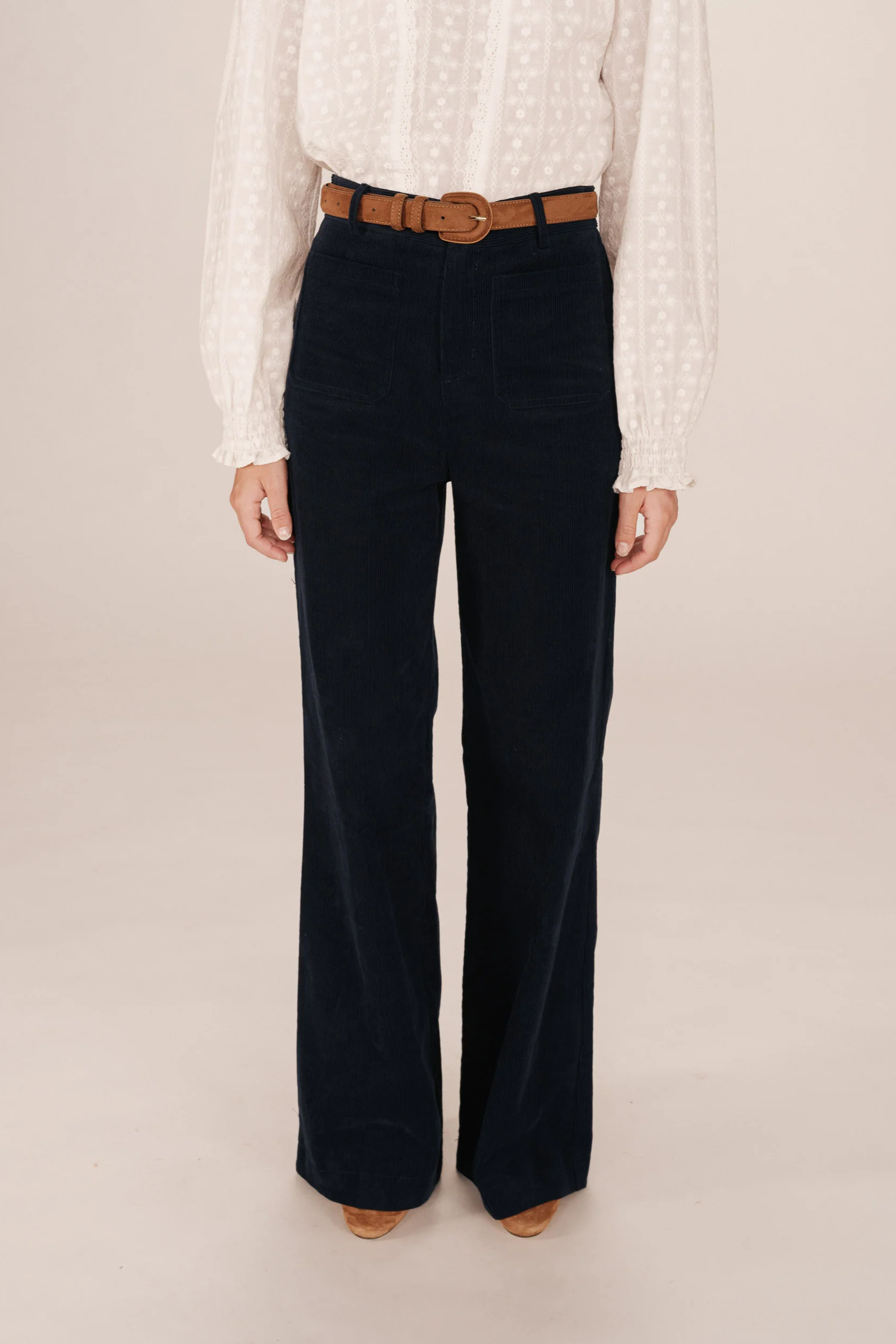 Person wearing a white blouse and dark blue velvet pants with a brown belt on a beige background
