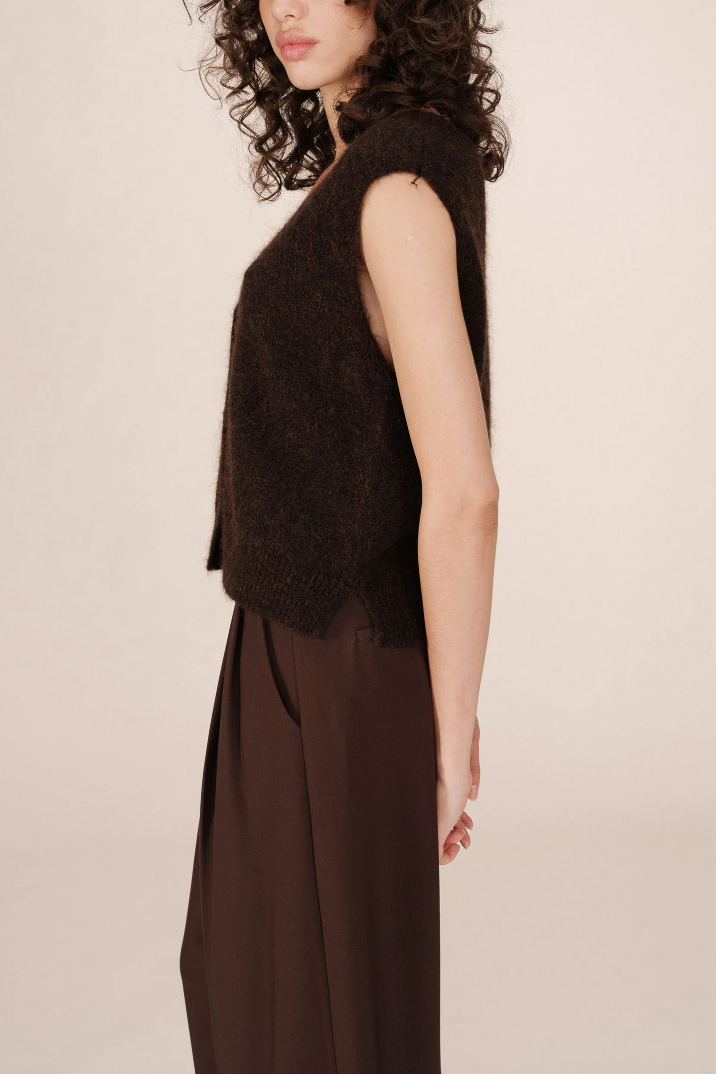Woman wearing a dark brown sleeveless top and dark pants against a light pink background