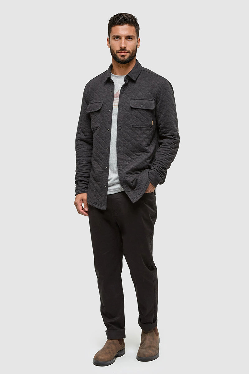 Man wearing a dark gray quilted jacket over a white shirt, black pants, and brown boots on a light gray background.
