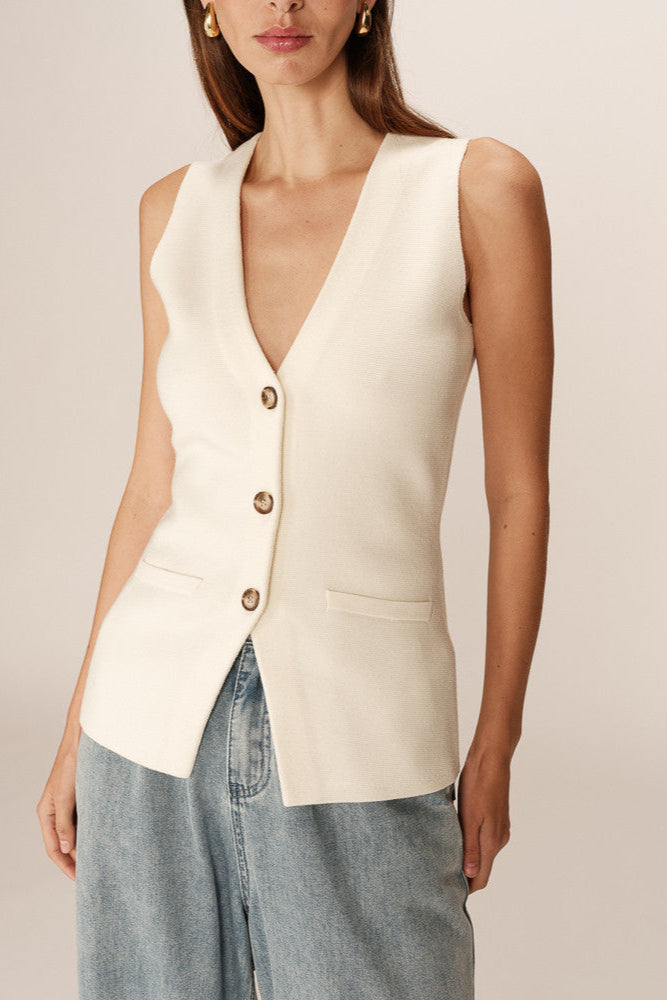 Woman wearing a cream-colored vest over a beige top with blue jeans on a plain background