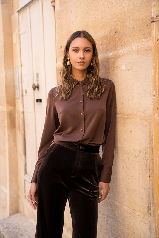 Woman wearing a brown blouse and black pants standing against a stone wall.