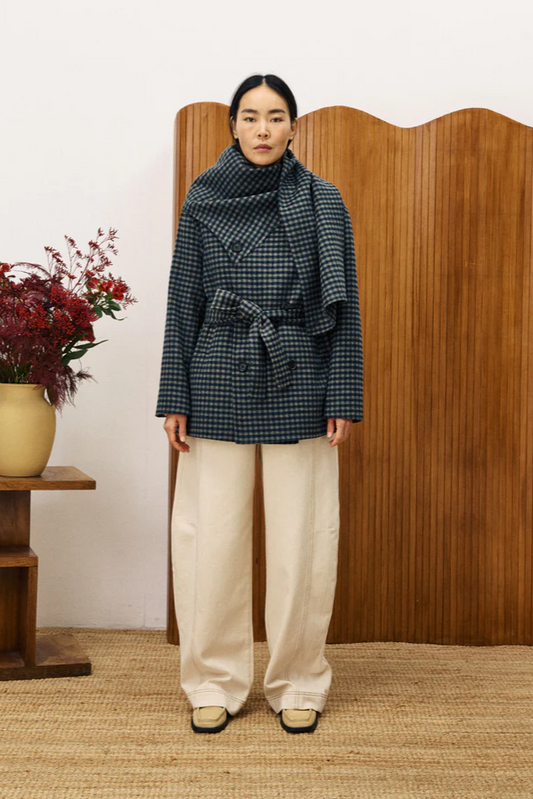 Person wearing a checkered coat and white pants standing in front of a wooden screen with a vase of flowers on a table.