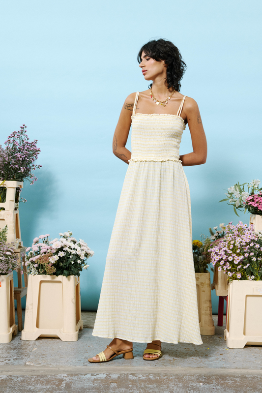 Woman in a butter yellow dress standing among potted flowers against a blue wall