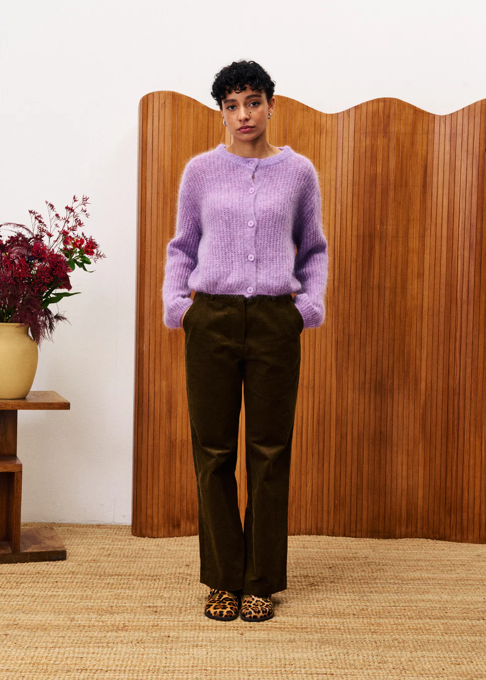 Person wearing a purple sweater and dark pants standing in front of a wooden screen with a decorative plant.