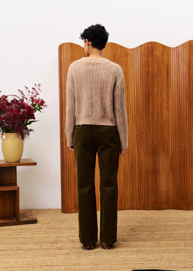 Person wearing a beige sweater and brown pants standing in front of a wooden screen with a vase of flowers on a table to their left.