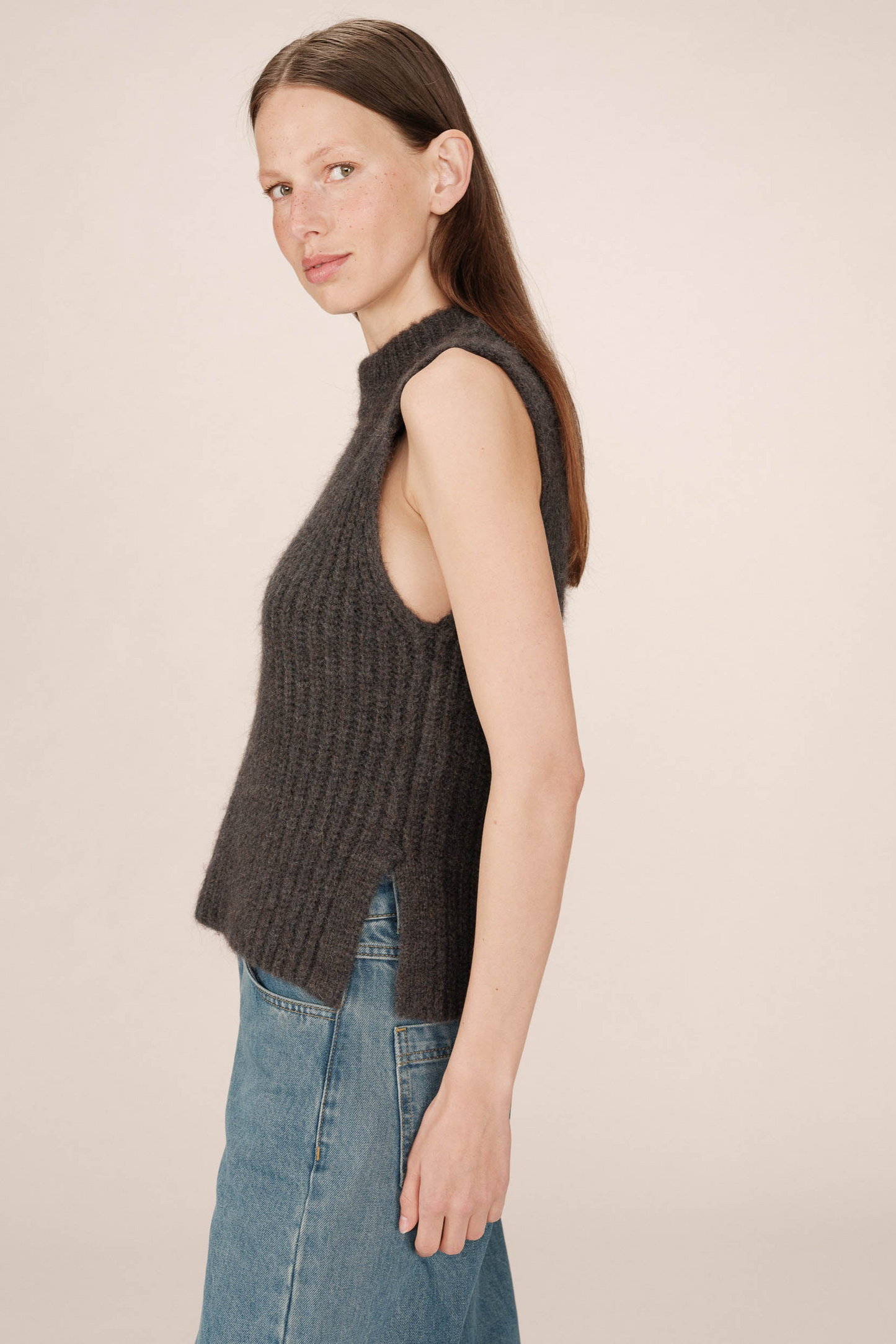Woman wearing a dark gray ribbed sleeveless sweater and blue jeans on a beige background