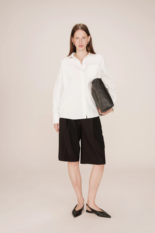 Woman wearing a white shirt and black shorts holding a black clutch against a plain background
