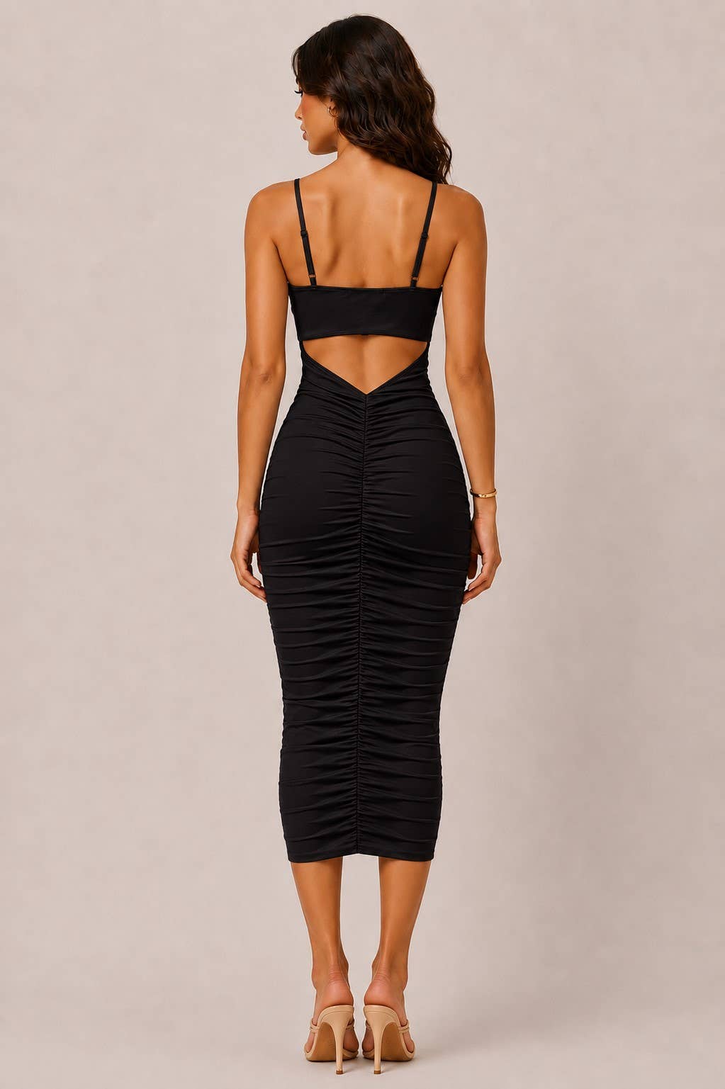 A woman shown from the back wearing a black ruched midi dress with spaghetti straps and a cutout at the upper back, paired with beige high-heeled sandals against a plain beige studio background.